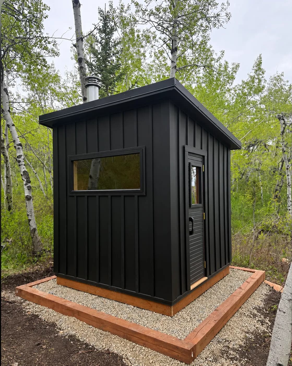Billy Oley’s black board-and-batten Model T with a wood-stove chimney, on a gravel pad among birch trees