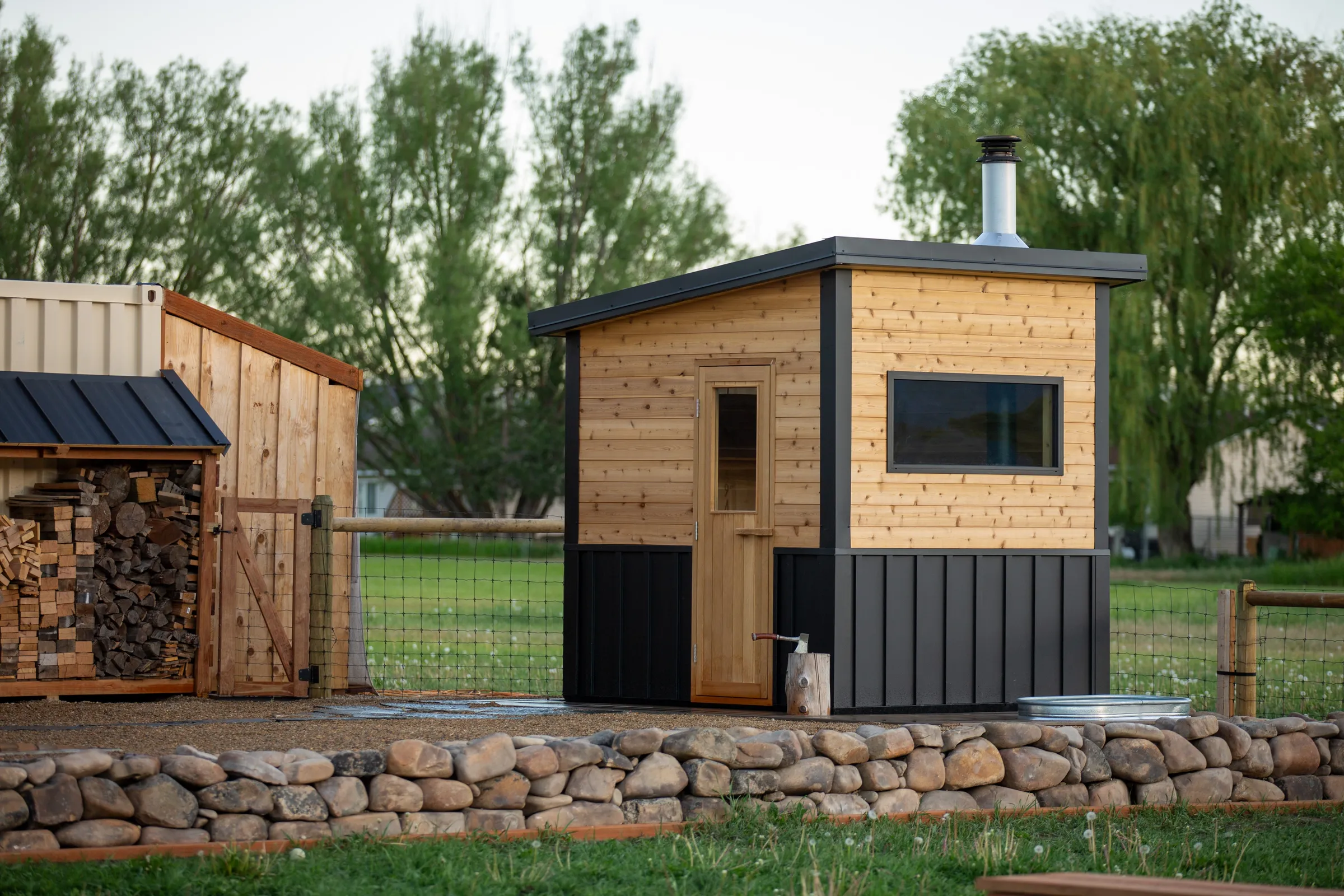 Model T with cedar and black metal siding in a fenced yard beside a woodpile