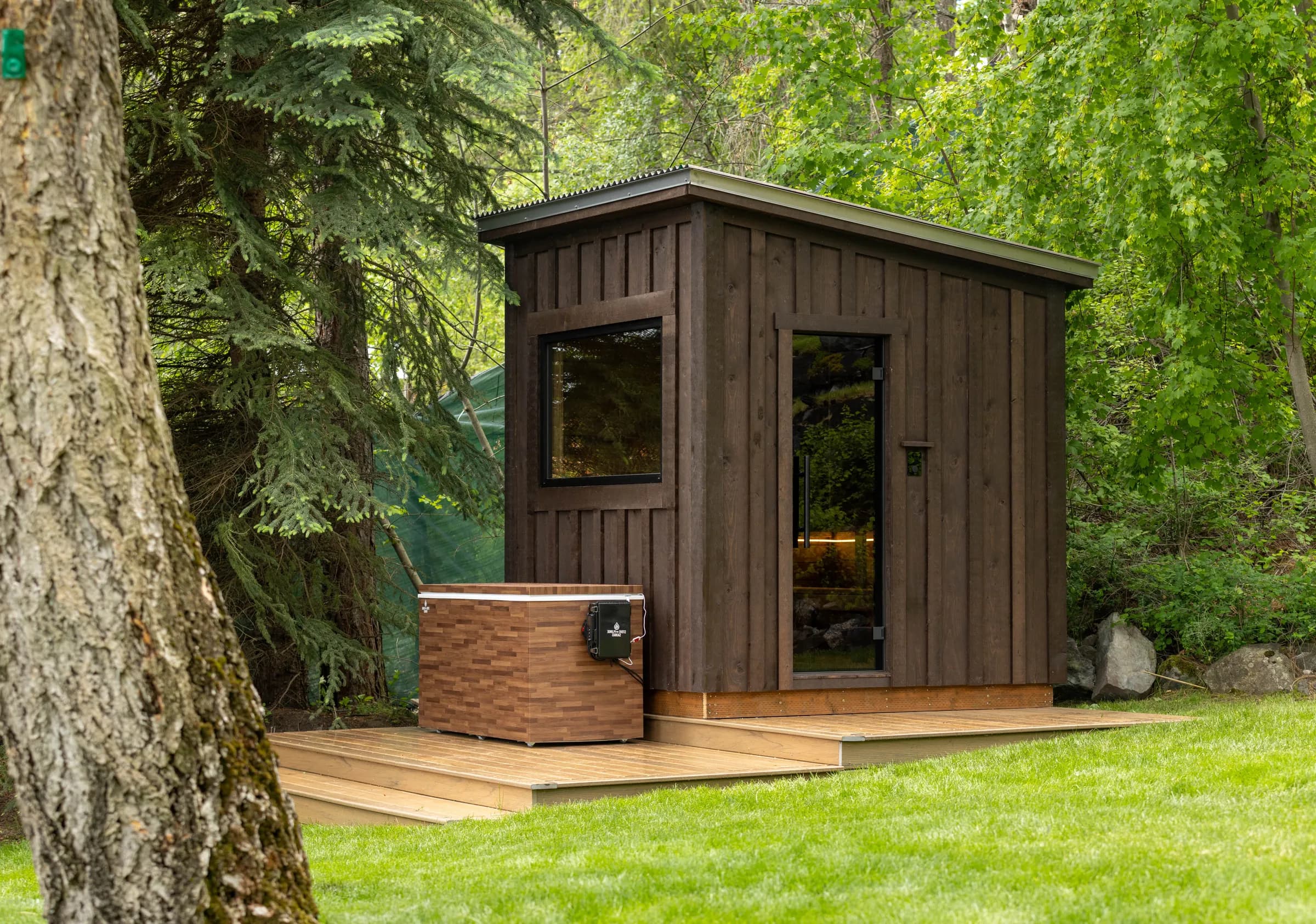 Dark-stained Model T sauna and a wood-clad cold plunge on a deck among trees