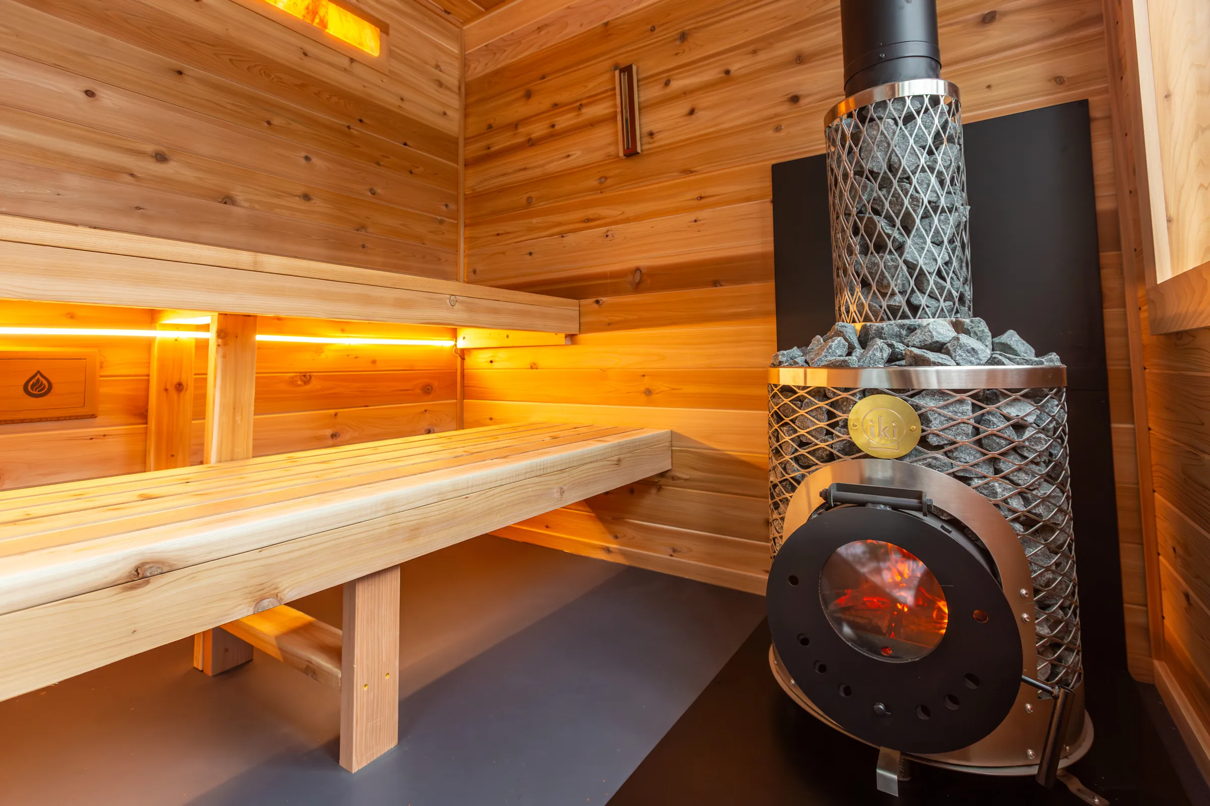 Wood-fired stove with a glass fire door beside cedar benches lit from below